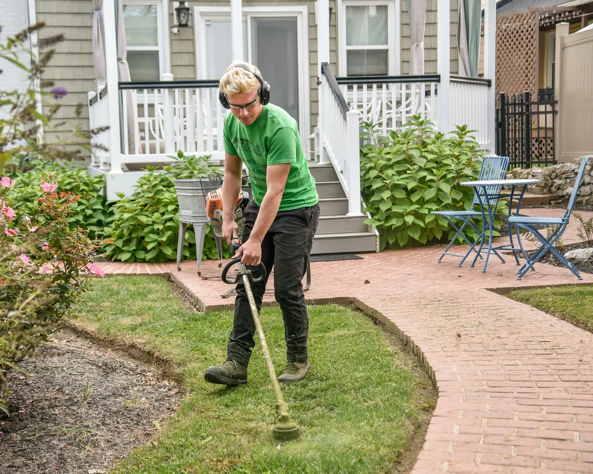 Un jardinier au travail devant une maison, coupe-bordure à la main.